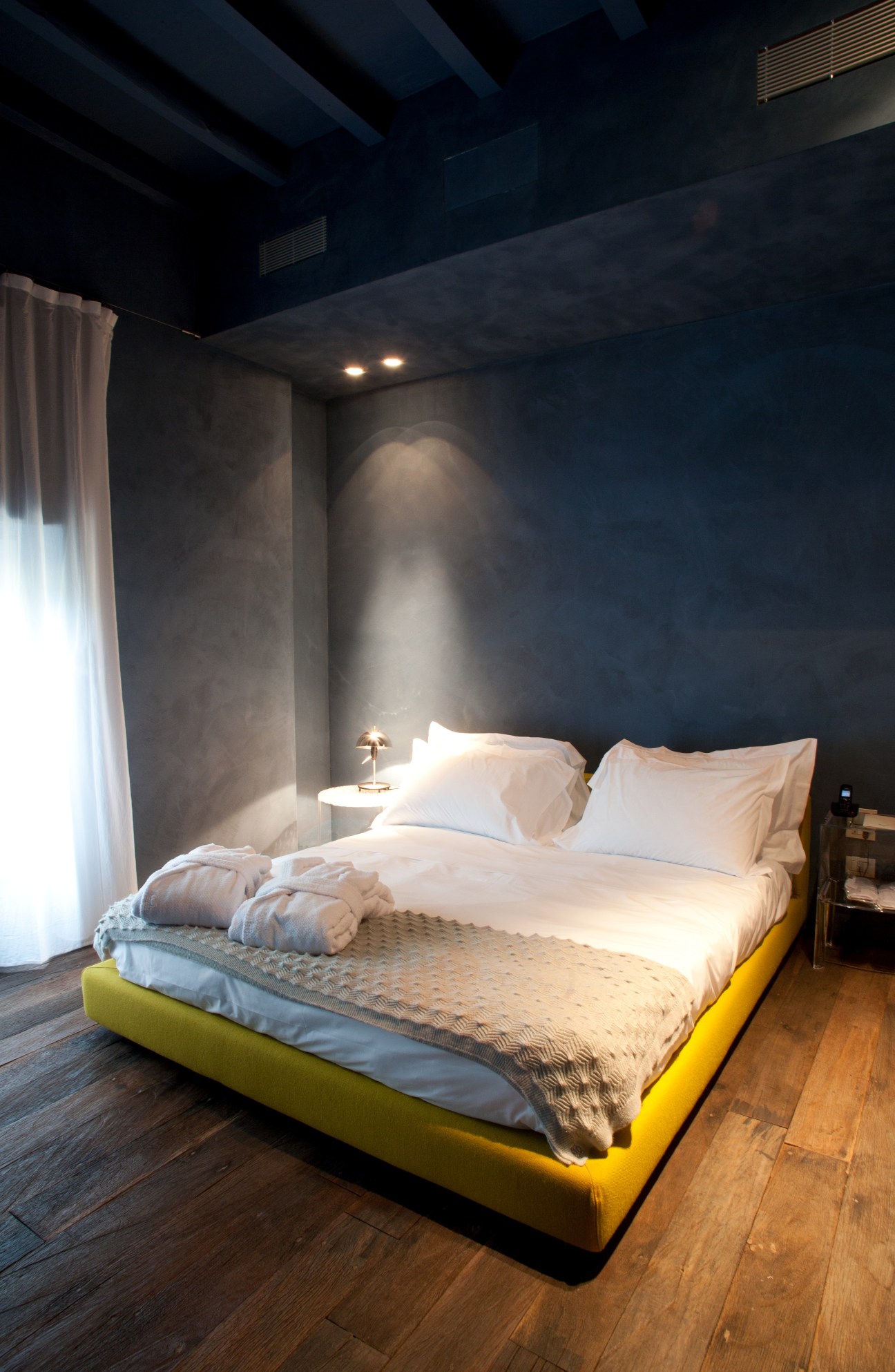Modern hotel bedroom with yellow bed frame, white linens, dark blue walls, and recessed lighting.