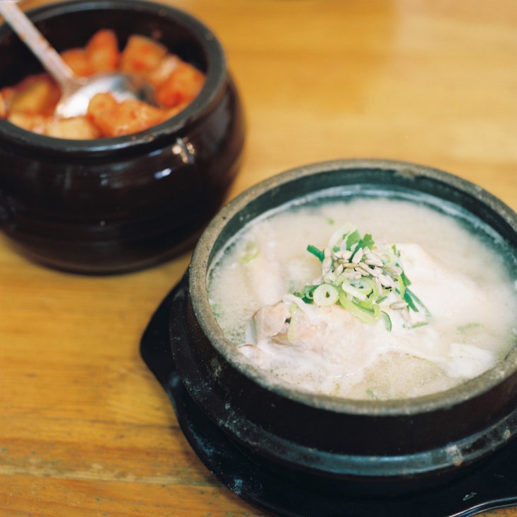 Bowl of samgyetang (ginseng chicken soup) with sliced green onions, served in a traditional stone bowl on a wooden table.