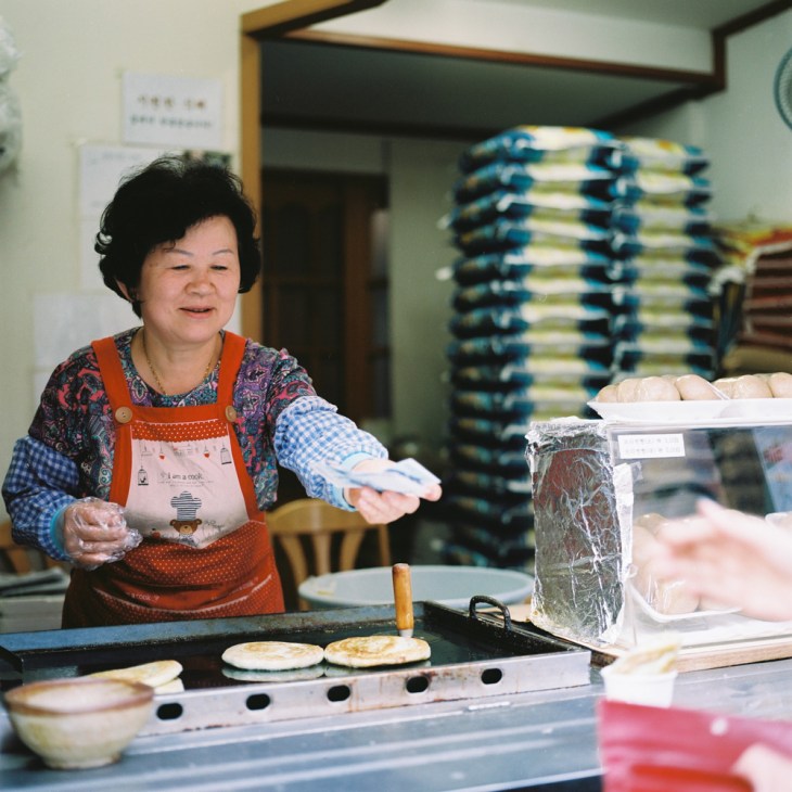 Woman in orange apron making hotteok rice pancakes at a street food stall in Seoul.