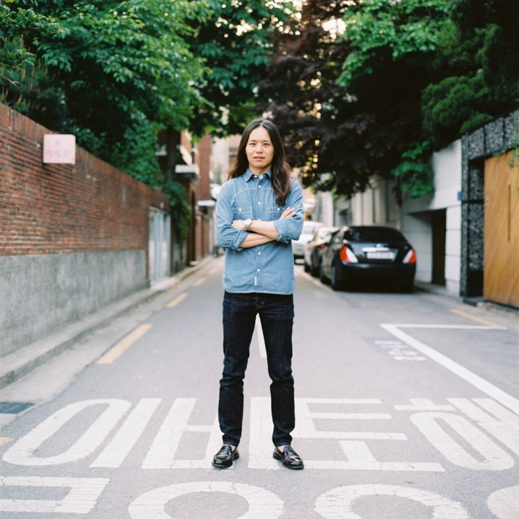 Covernat director Yun Hyungseok stands on a crosswalk in a tree-lined alley wearing a denim shirt and dark jeans.