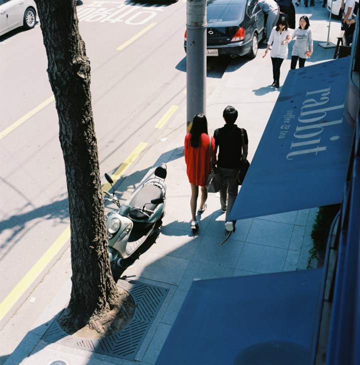 Overhead view of a woman in a red coat walking with a man in black on a sunny Seoul street with a scooter and tree.