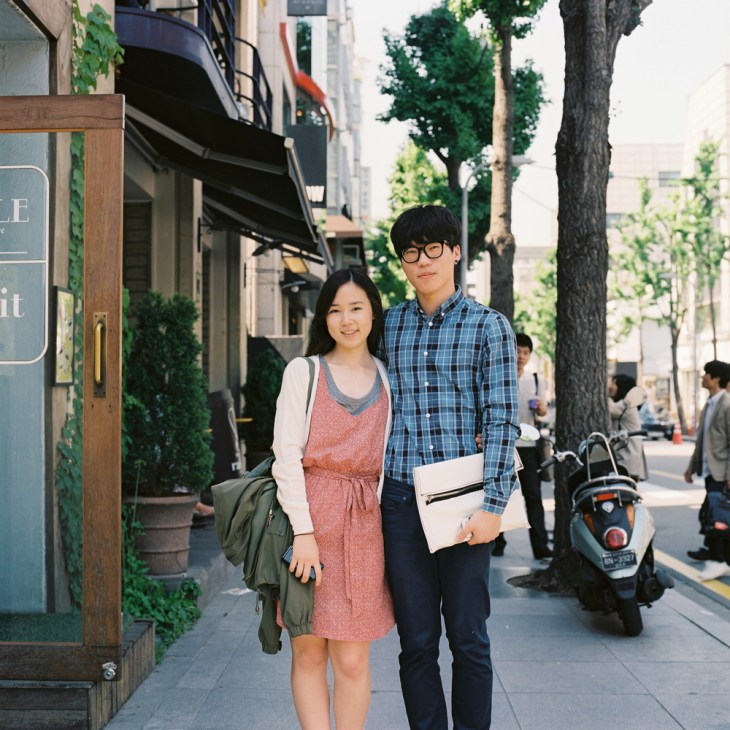 Young university student couple standing on a tree-lined Seoul street with shops and buildings behind them.