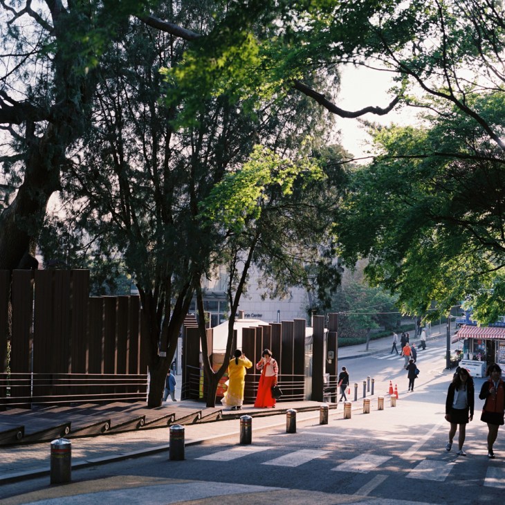Pedestrians walk through a shaded tree-lined street in Seoul's Samcheong-dong neighborhood with modern buildings visible in the background.