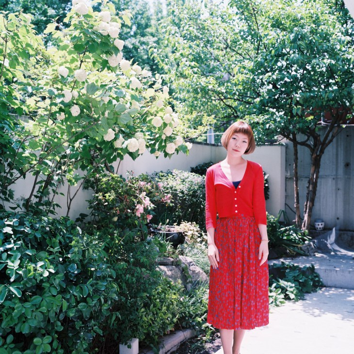 Kim Gayoung, designer at Jardin de Chouette, wearing a red cardigan and pleated skirt in a lush garden setting with white flowering shrubs.