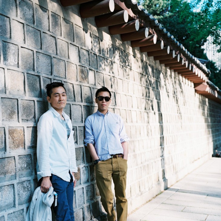 Two men standing against a traditional Korean stone wall with decorative roof eaves, one in white shirt and one in blue shirt.
