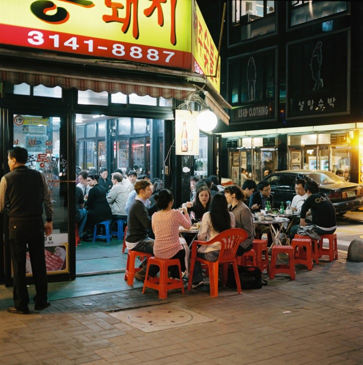 Diners seated at red plastic chairs outside a Korean street food vendor in Hongdae, Seoul, at night.