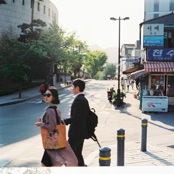 Two people walking on a sunny Seoul street with traditional buildings, shops, and signage in Samcheong-dong.