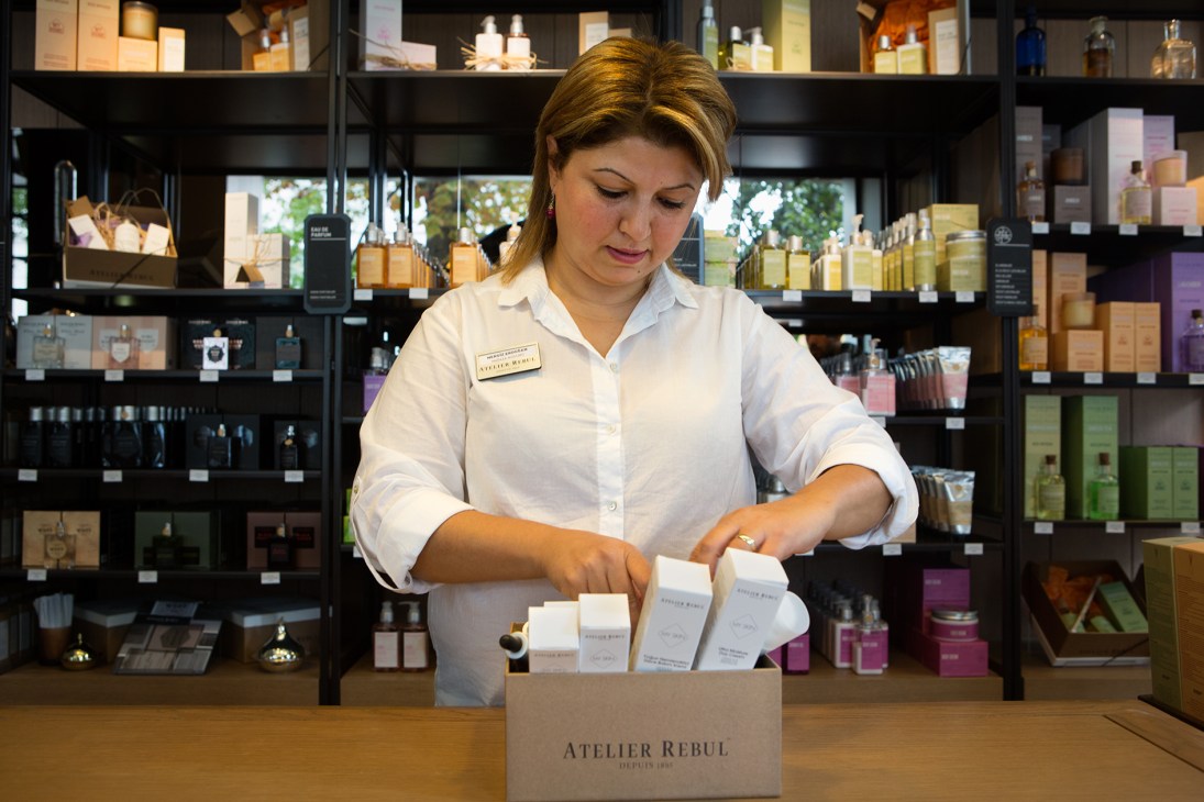 Staff member at Atelier Rebul boutique arranging fragrance products at counter with shelves of perfumes behind.