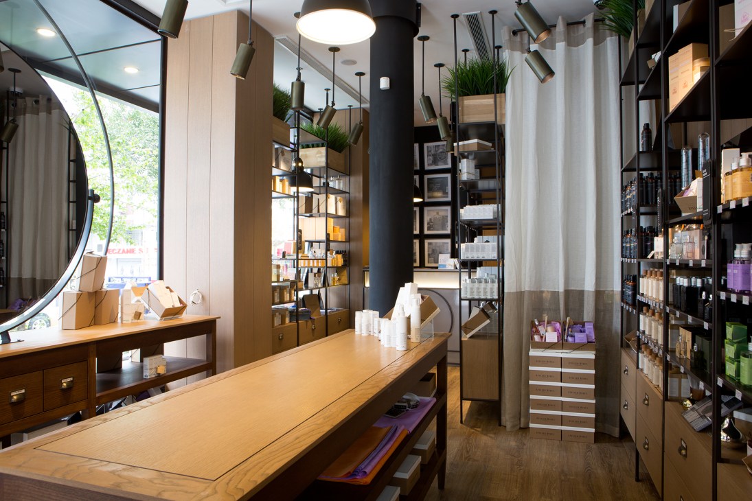 Interior of Atelier Rebul boutique featuring wooden counter, shelving with fragrance products, pendant lighting, and large curved mirror.