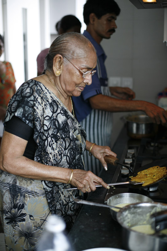 Subodh Gupta's mother cooking in the kitchen, wearing glasses and a patterned black shirt.