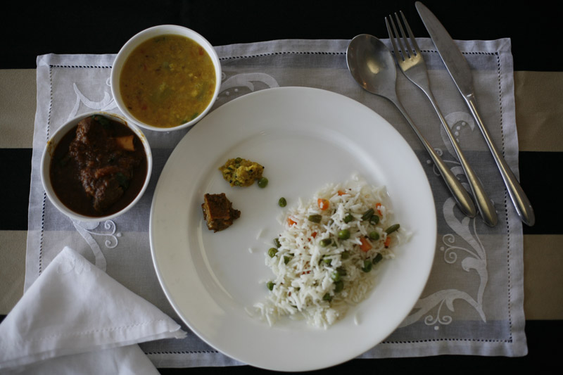 Overhead view of a plated meal with white rice mixed with peas and carrots, lamb curry in a bowl, and pakoras on a striped placemat with cutlery.