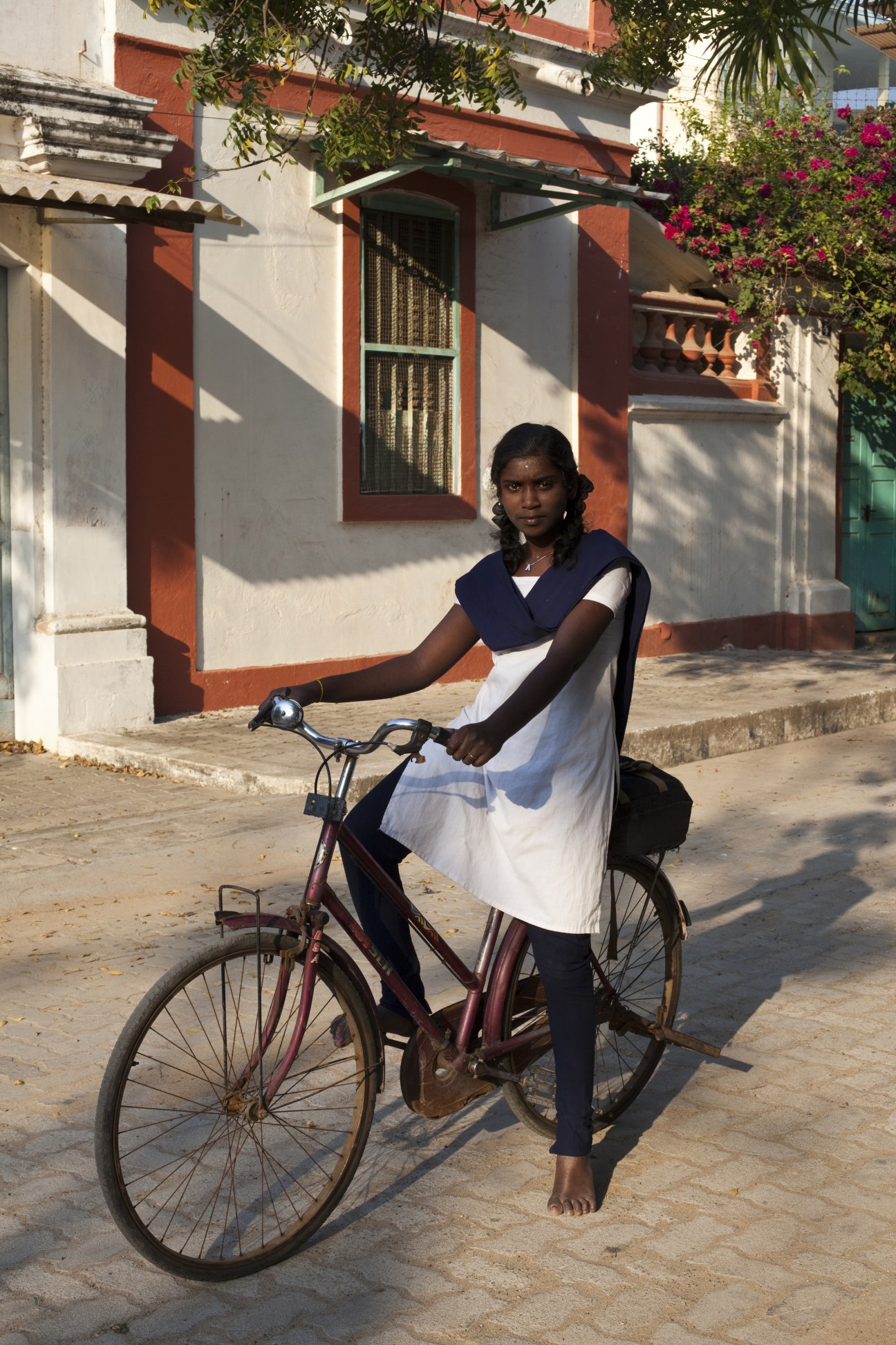 A young schoolgirl in uniform poses with a bicycle in front of a colonial-style building in Pondicherry.