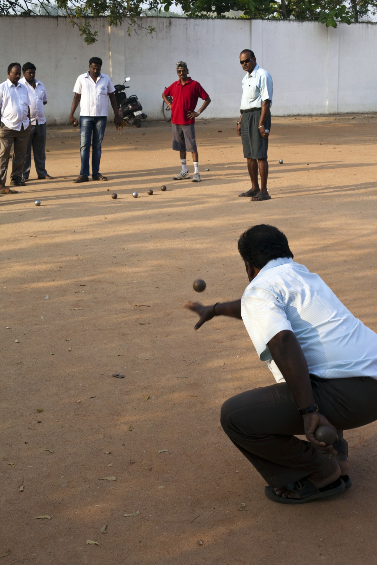 Men playing pétanque on a red-earth field in Pondicherry, with spectators watching nearby.