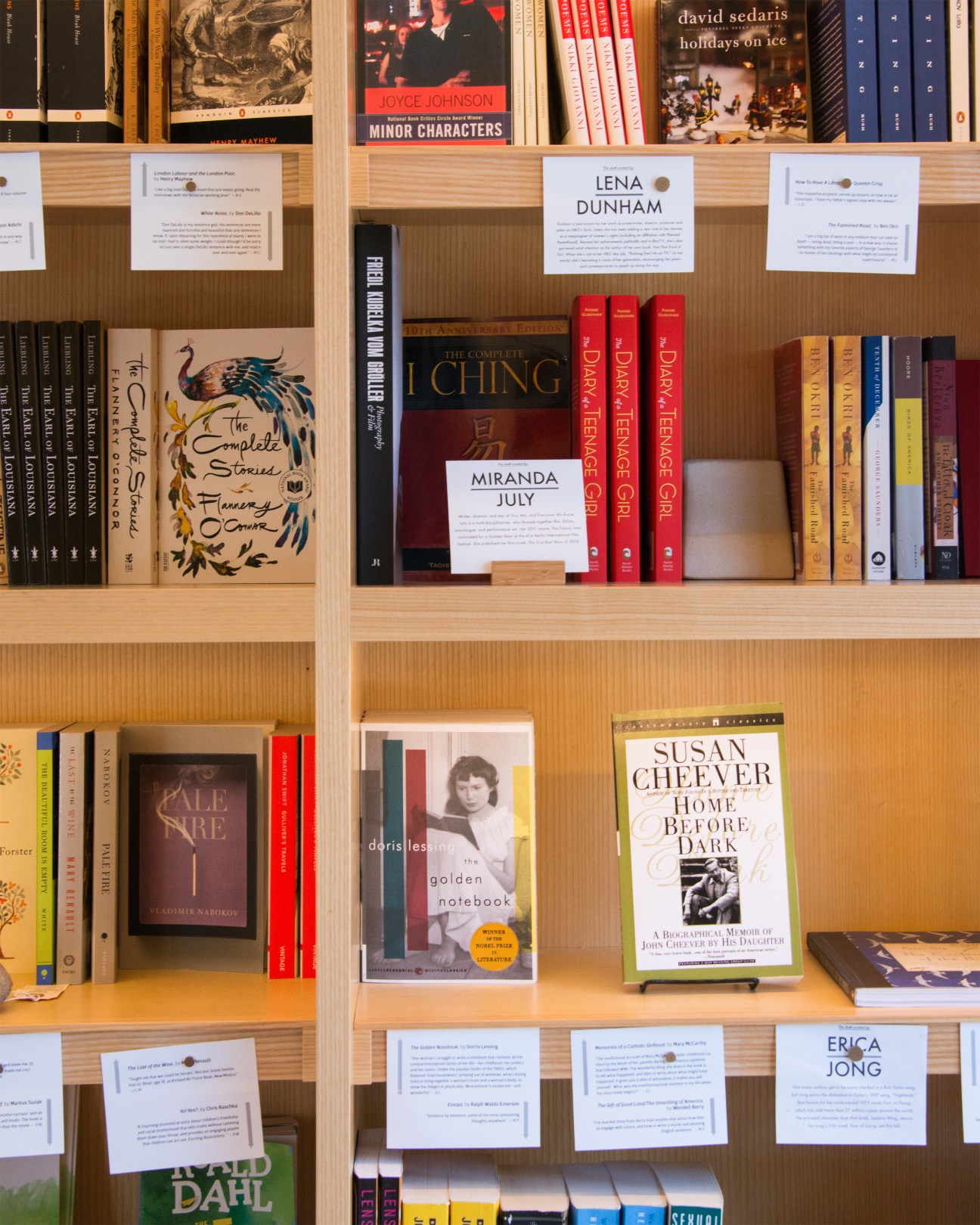 Wooden bookshelves at One Grand displaying guest-curated book collections with curator name cards.
