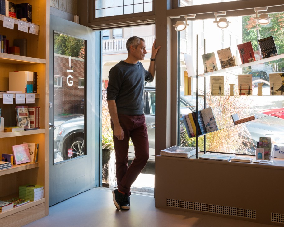 Aaron Hicklin stands in One Grand bookshop, browsing shelves of curated books with large windows behind him.