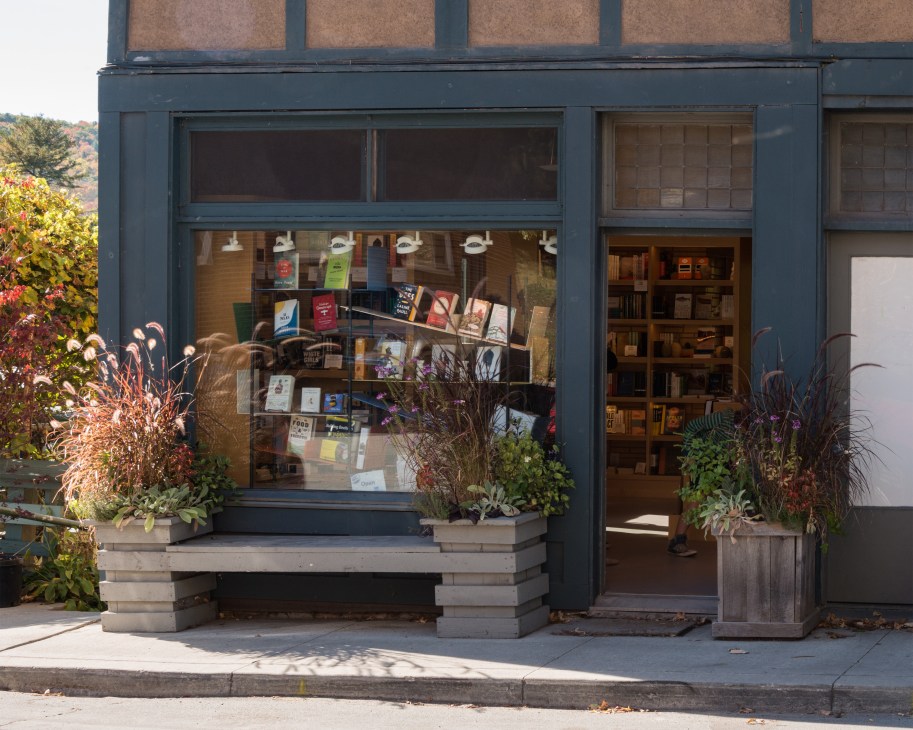 One Grand bookshop storefront with dark-framed windows displaying books and planters with autumn foliage.