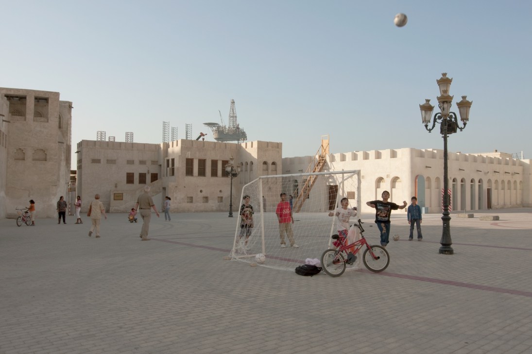 A football pitch with nets installed in a desert courtyard with traditional architecture and oil derricks visible in the background.