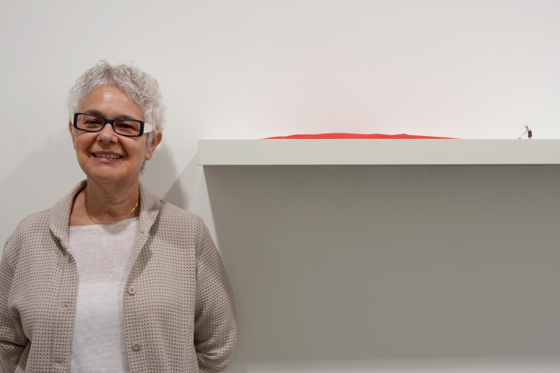 Woman with white hair and glasses smiling next to Liliana Porter's minimalist artwork featuring a red line on a grey surface.