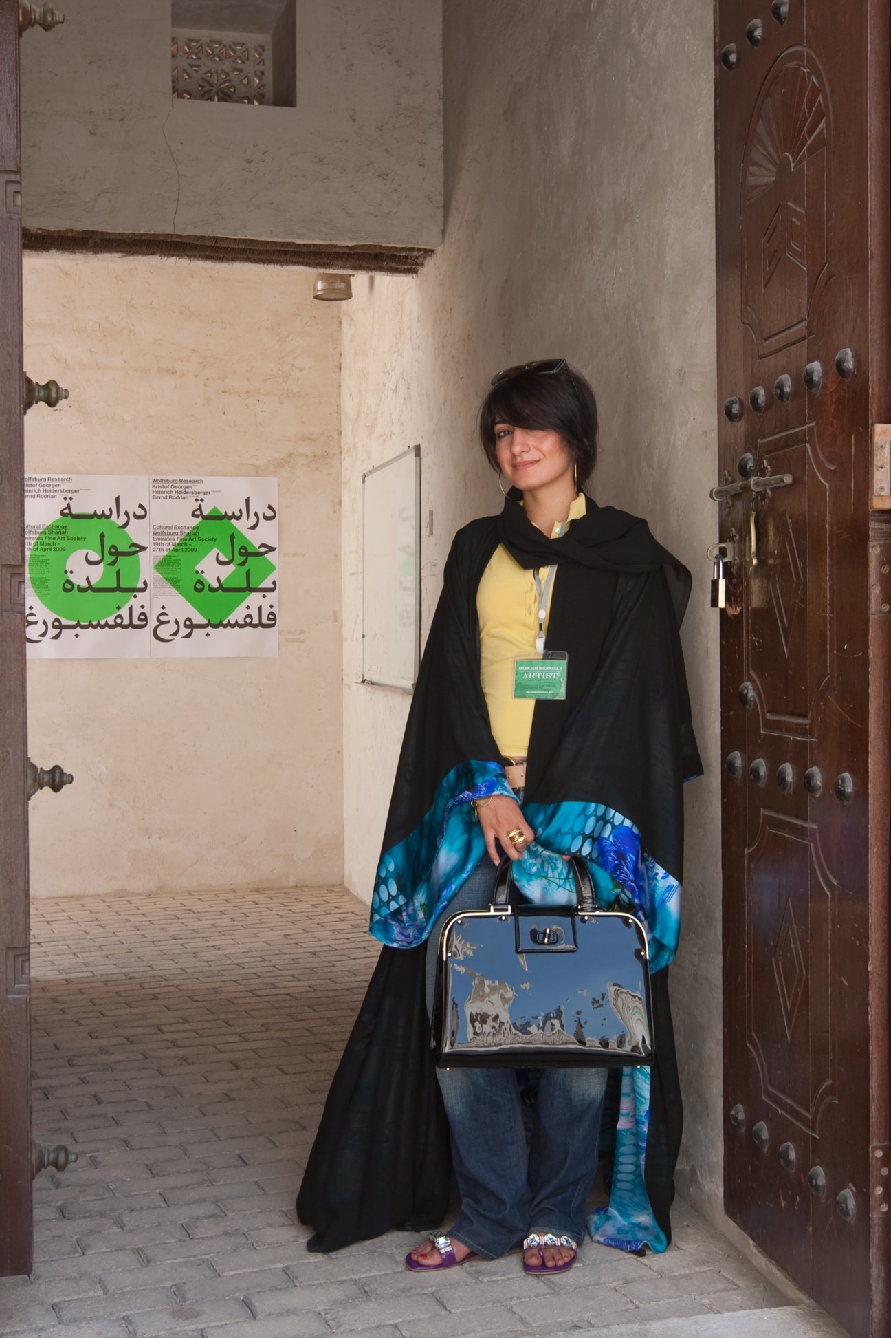 Lamya Gargash stands in a Sharjah alleyway wearing an abaya, yellow shirt, and holding a reflective briefcase.