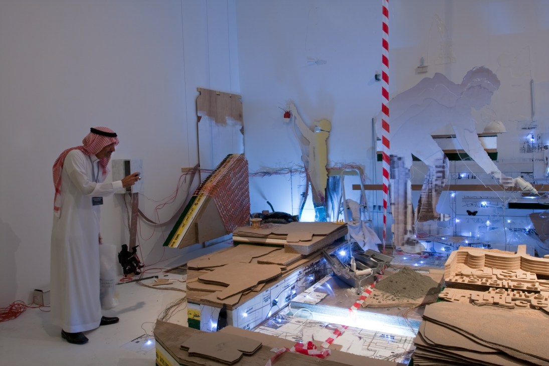 An Emirati man in traditional white thobe photographs Reem Al Ghaith's sculptural installation made of chipboard, sand, and construction materials.