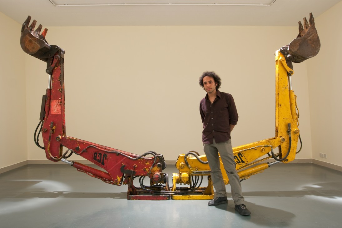 Artist Nida Sinnokrot stands between two large industrial excavator arms painted red and yellow in a gallery space.