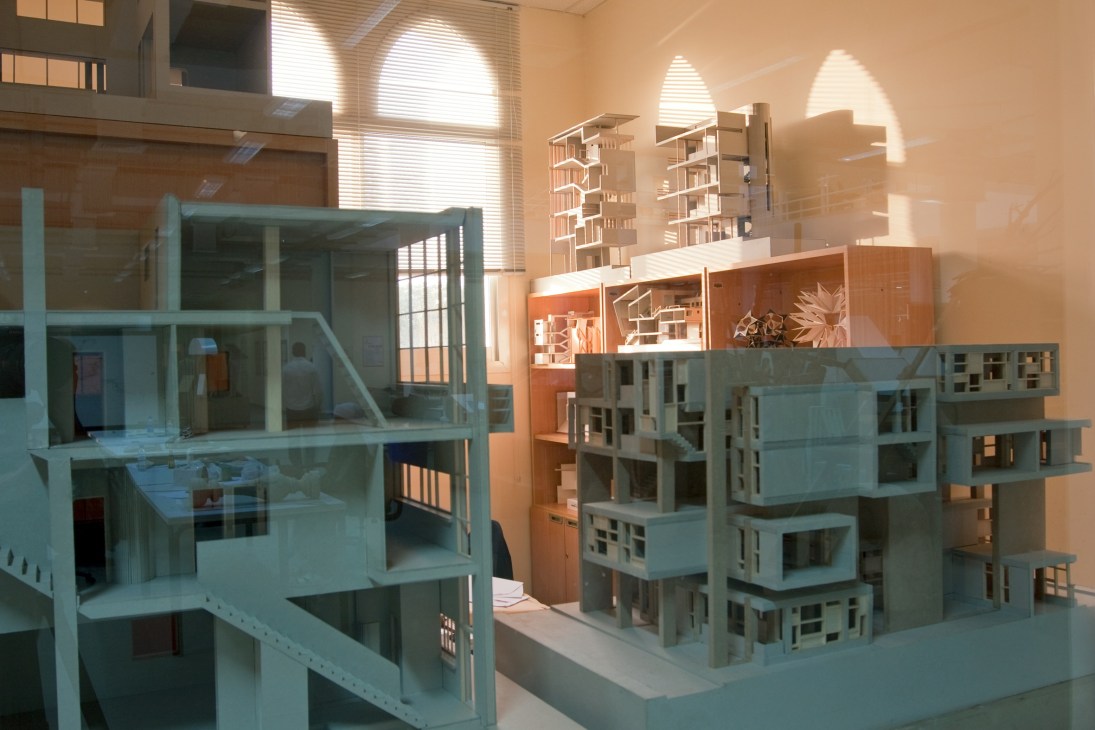 Architectural model buildings in blue and orange displayed on white platforms in a sunlit gallery space.