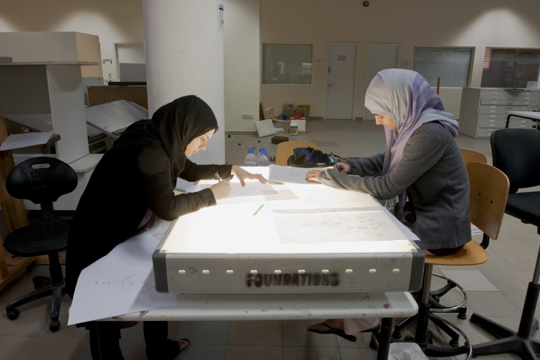 Two female design students wearing headscarves work at a light table in a studio classroom.