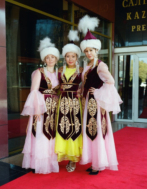 Three women wearing traditional Kazakh folk costumes with embroidered vests, pink layered dresses, white fur hats, and beaded accessories standing on a red carpet outside a building.