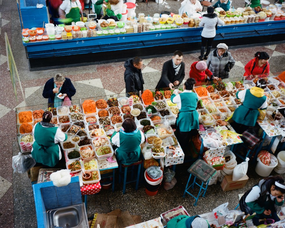 Overhead view of Almaty's Kok-Bazaar with vendors in teal uniforms selling colorful prepared foods to customers.