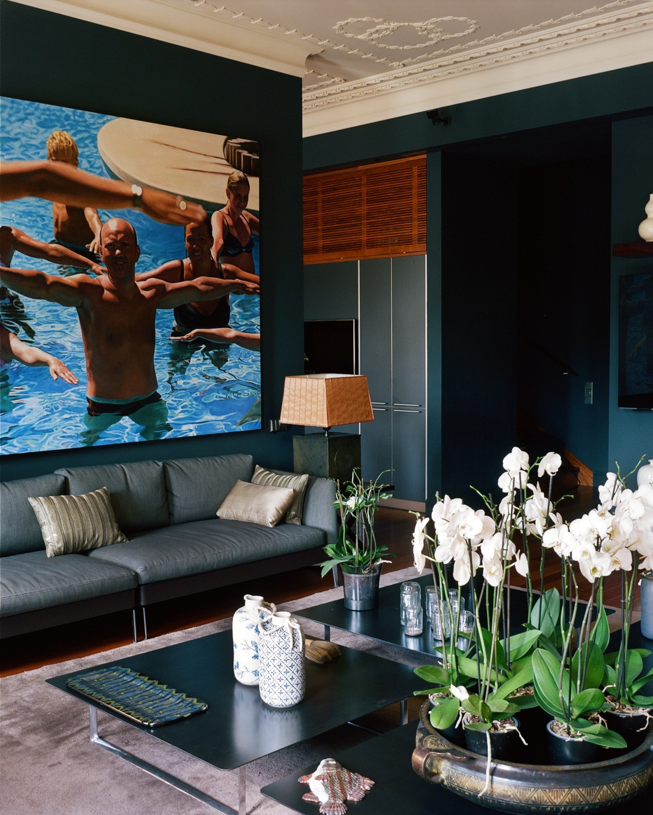Modern living room with dark teal walls, ornate ceiling, black sofa, white orchids, and contemporary art above.
