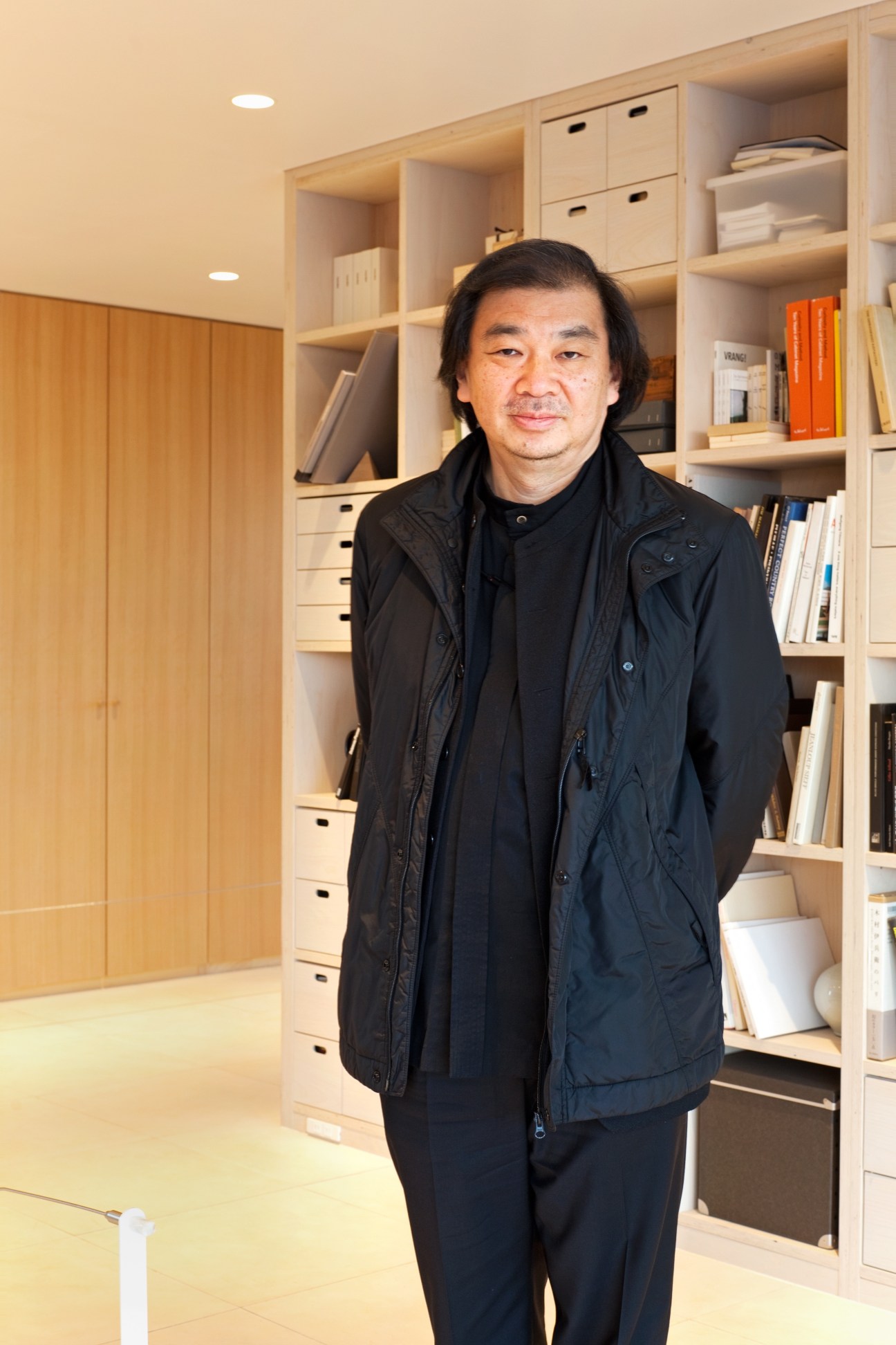 Architect Shigeru Ban standing in a modern interior with light wood shelving and storage units behind him.