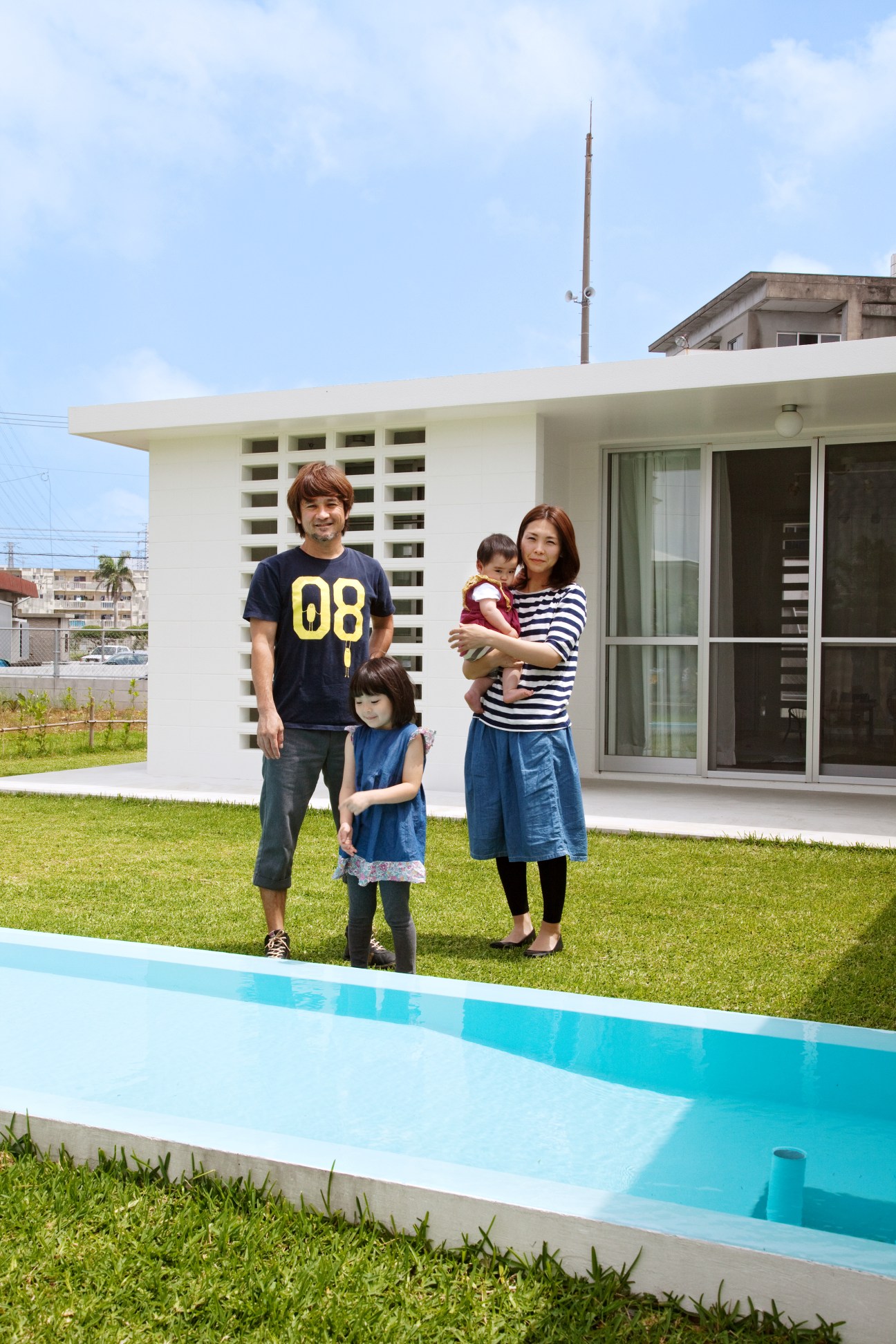 Hairdresser Shigeo Shima with his wife Ayumi and their children standing in front of their white Okinawa Standard house with a pool.