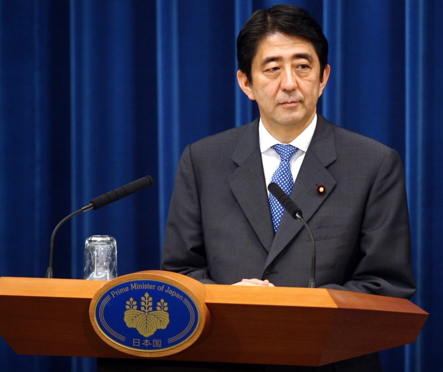 Shinzo Abe at a podium with the Prime Minister of Japan seal, wearing a dark suit and blue tie against a blue curtain background.
