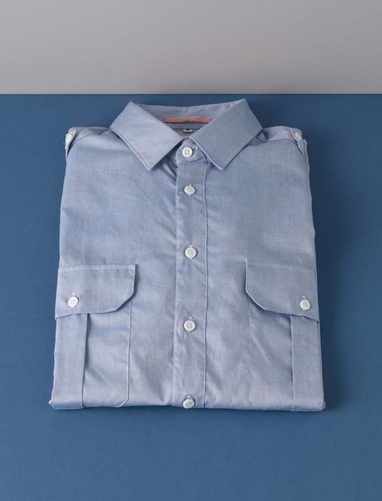 Light-blue Oxford shirt with military-style pocket detailing by Apolis Activism, laid flat on blue background.