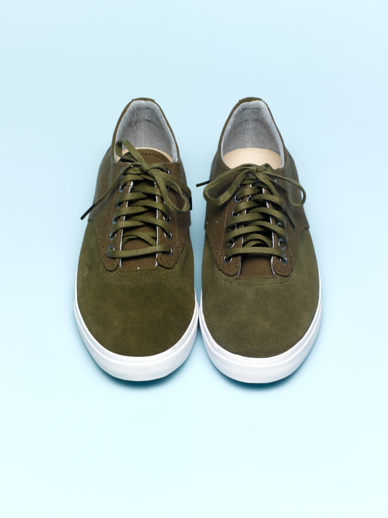Pair of olive green canvas lace-up sneakers with white sole and gray interior lining on light blue background.
