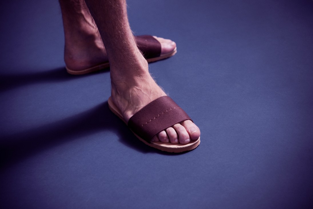 Person wearing burgundy house slippers with tan soles on a blue background.