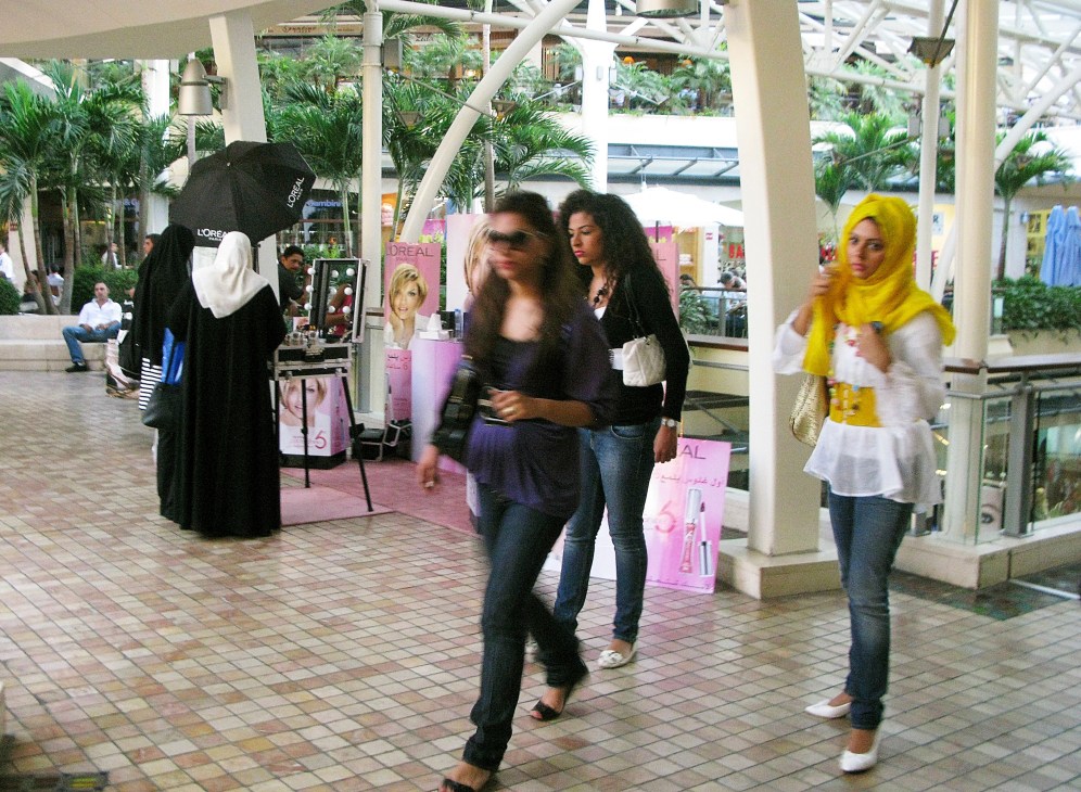 Shoppers walking through a modern mall with palm trees, storefronts and pink cosmetic brand signage.