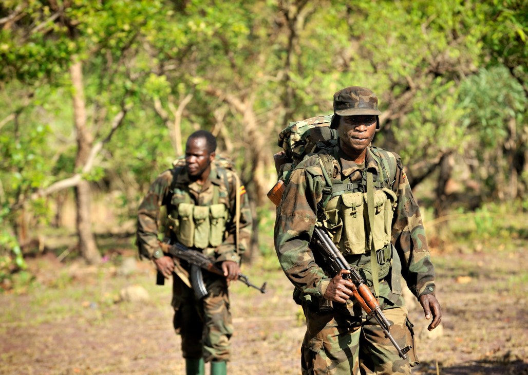 Bangladeshi military soldiers in camouflage uniforms and carrying rifles walk through a forested area.