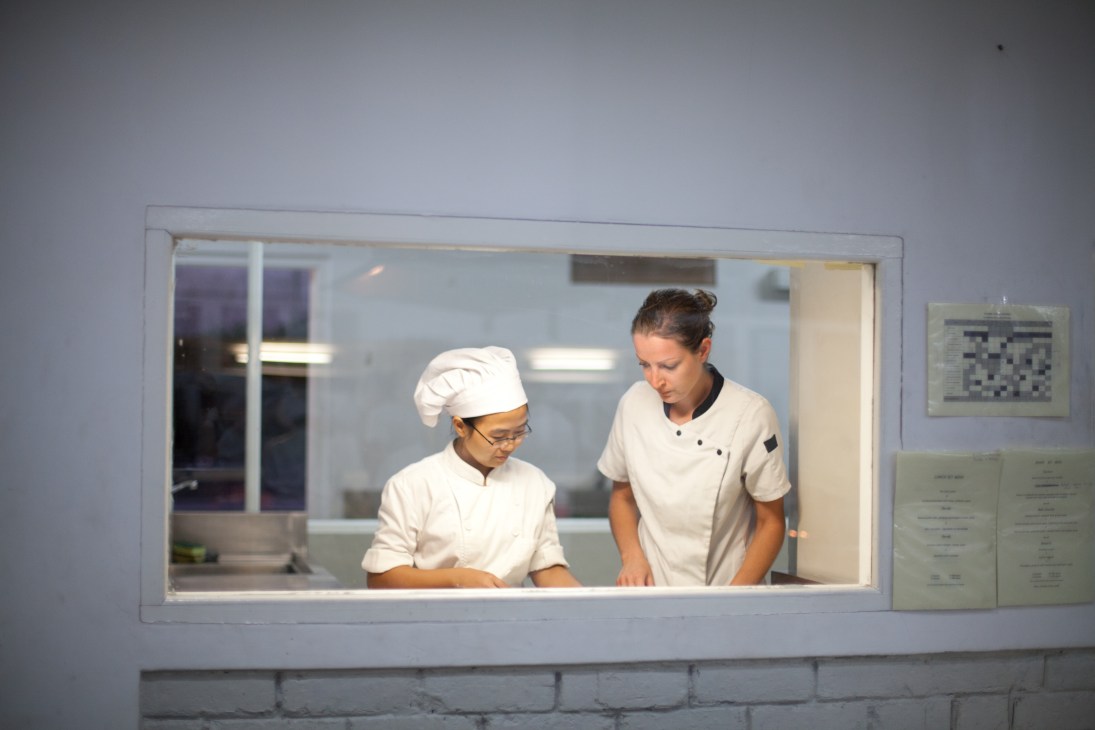 Two chefs in white uniforms and hat working together in a modern kitchen visible through a service window.