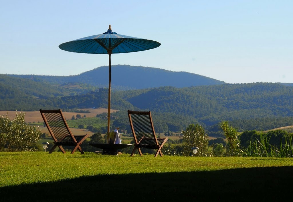 Two wooden lounge chairs beneath a blue umbrella overlook rolling green Tuscan hills.