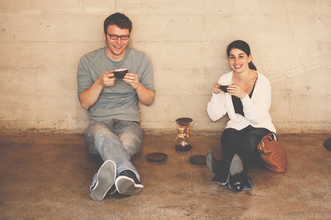 Two people sitting against a wall holding black cups and smiling while looking at mobile devices.