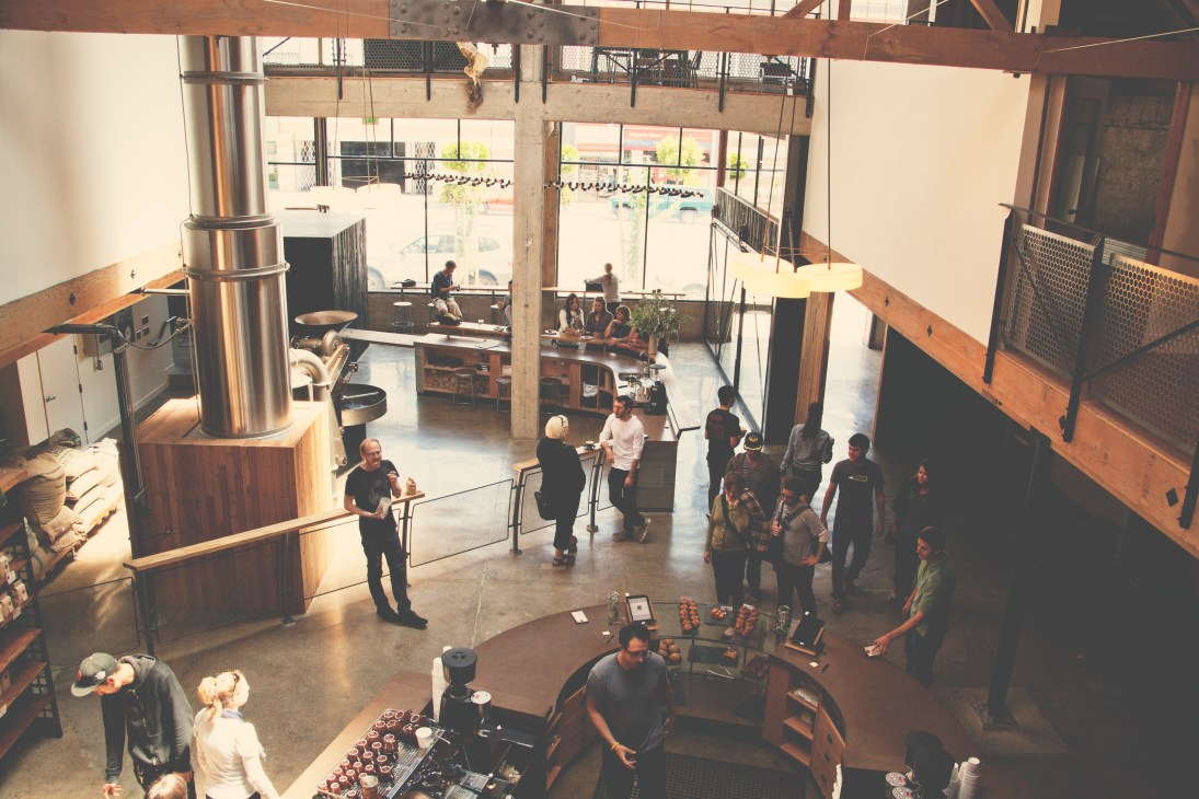 Interior of Sightglass coffee roastery in San Francisco showing customers and staff in a loft-style space with exposed beams and metal roasting equipment.
