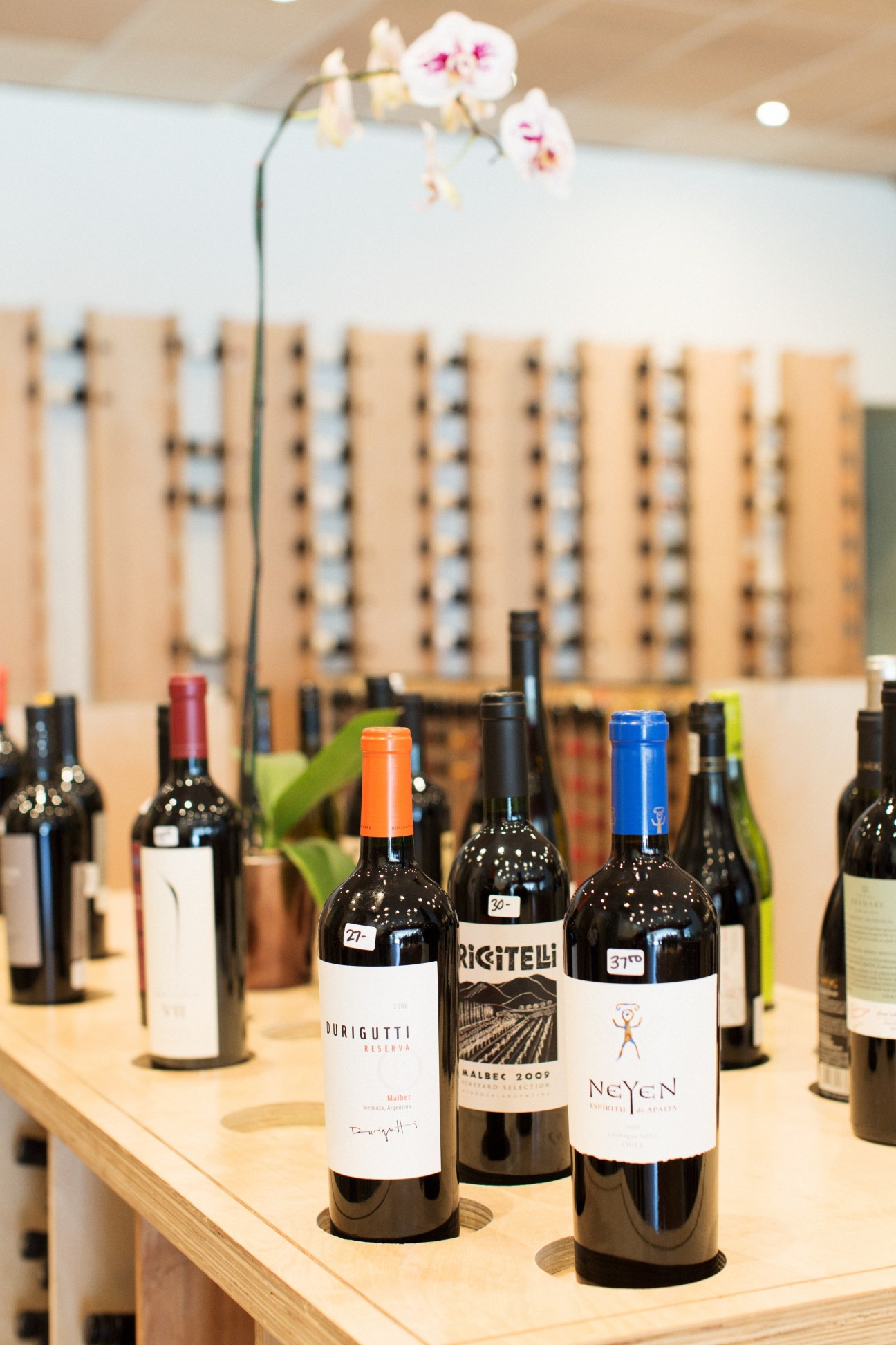 Wine bottles displayed on wooden counter with orchid flowers and wooden slat wall backdrop.