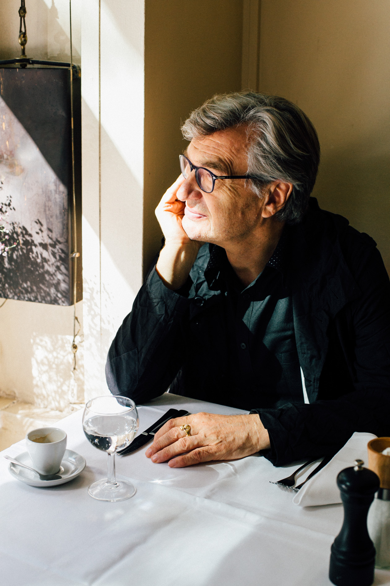 Wim Wenders sits at a restaurant table with an espresso cup, glass of water, and a framed photograph on the wall behind him.