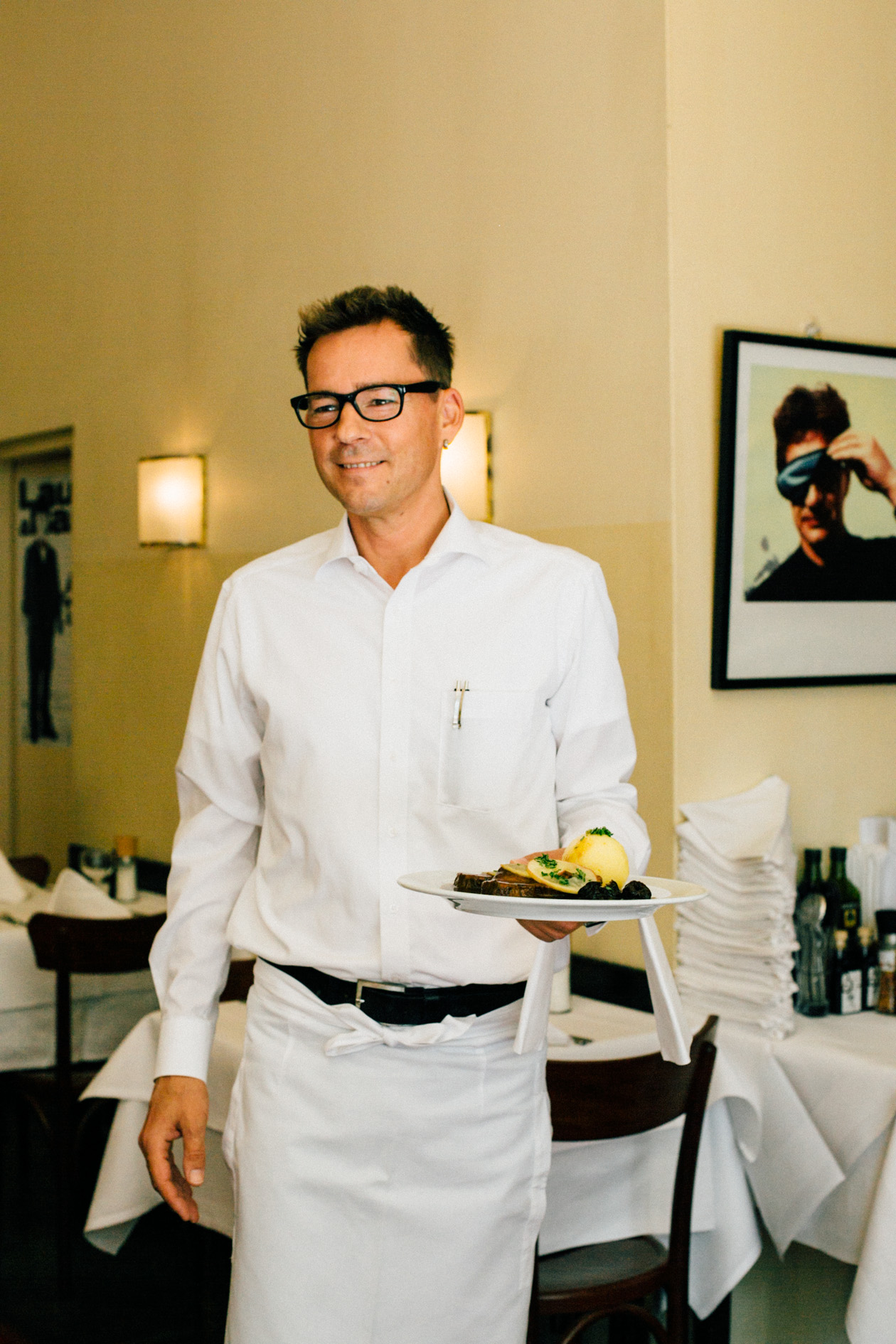 Waiter in white shirt and apron serving a plate with fish and potatoes in a restaurant dining room.