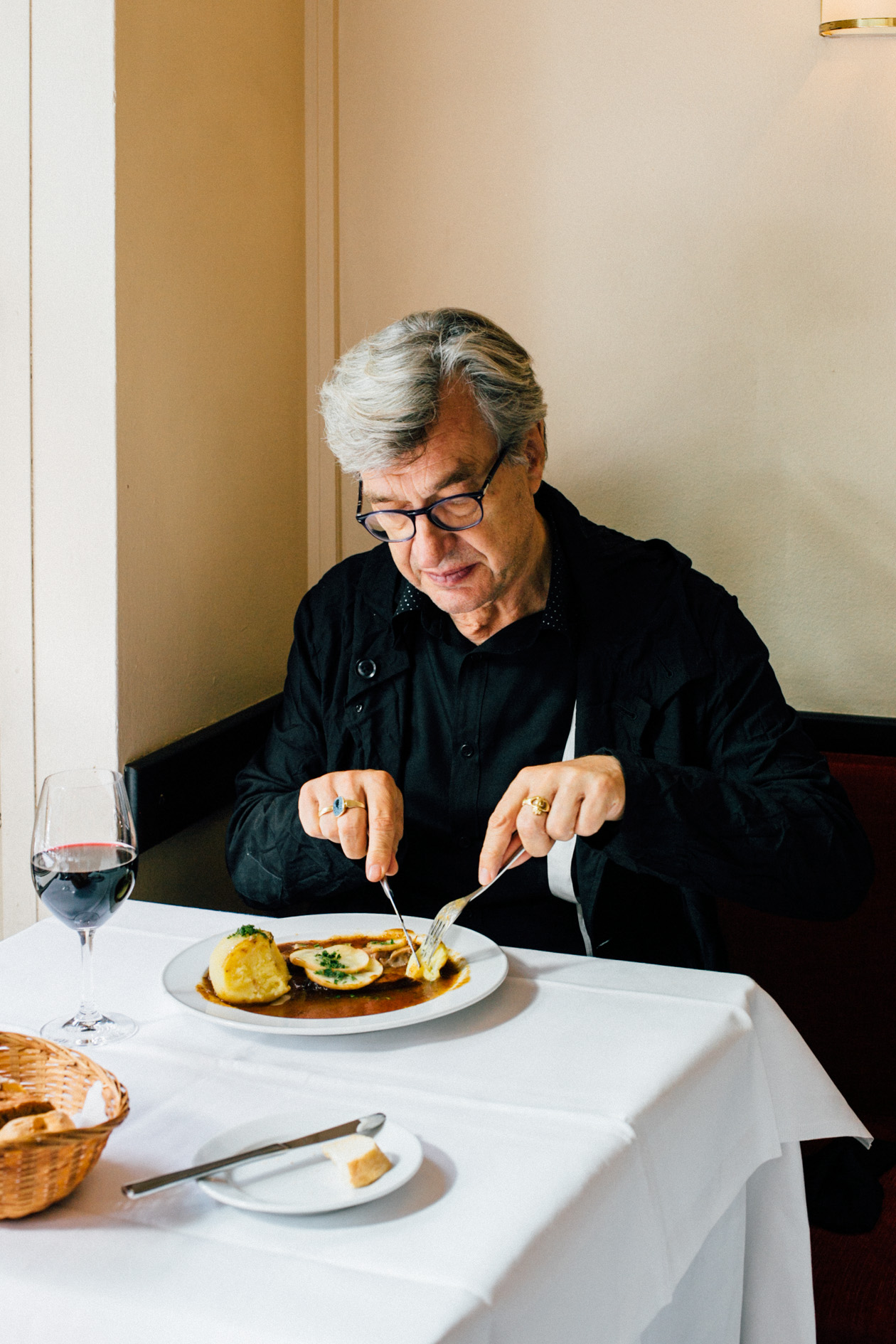 Wim Wenders eating pork roast at restaurant table with wine glass and bread basket.