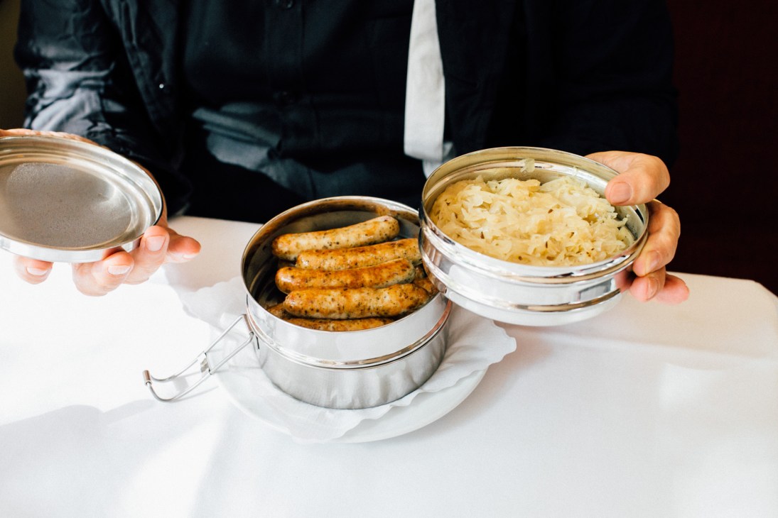 Hands holding open metal lunchbox containers with sausages and sauerkraut.