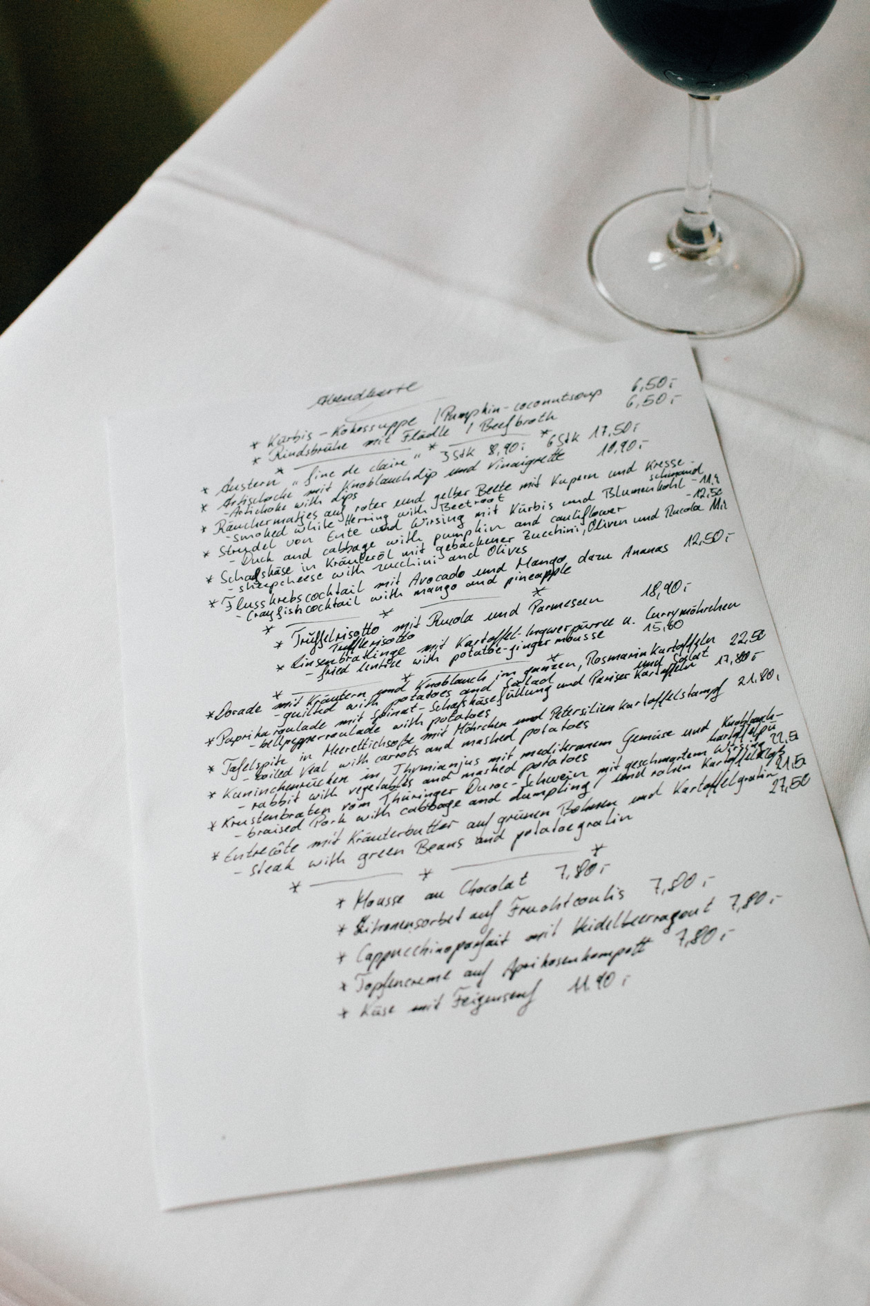 Handwritten restaurant menu on white paper next to a glass of red wine on a table.