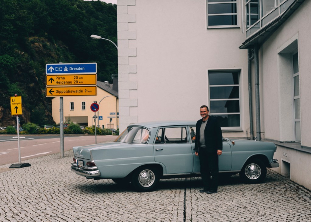 Man in dark blazer standing beside a light blue vintage car near German road signs in Glashütte.