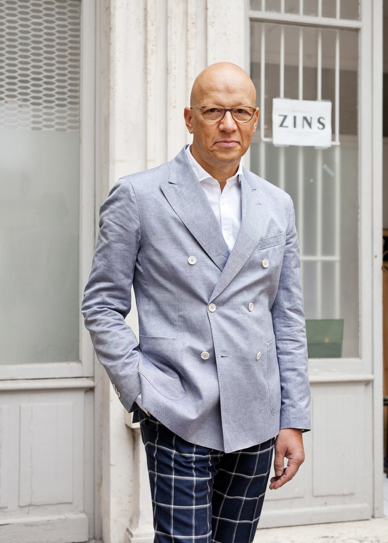 Frank Zins, a man wearing glasses and a light blue double-breasted blazer, stands outside a storefront with a 'ZINS' sign.